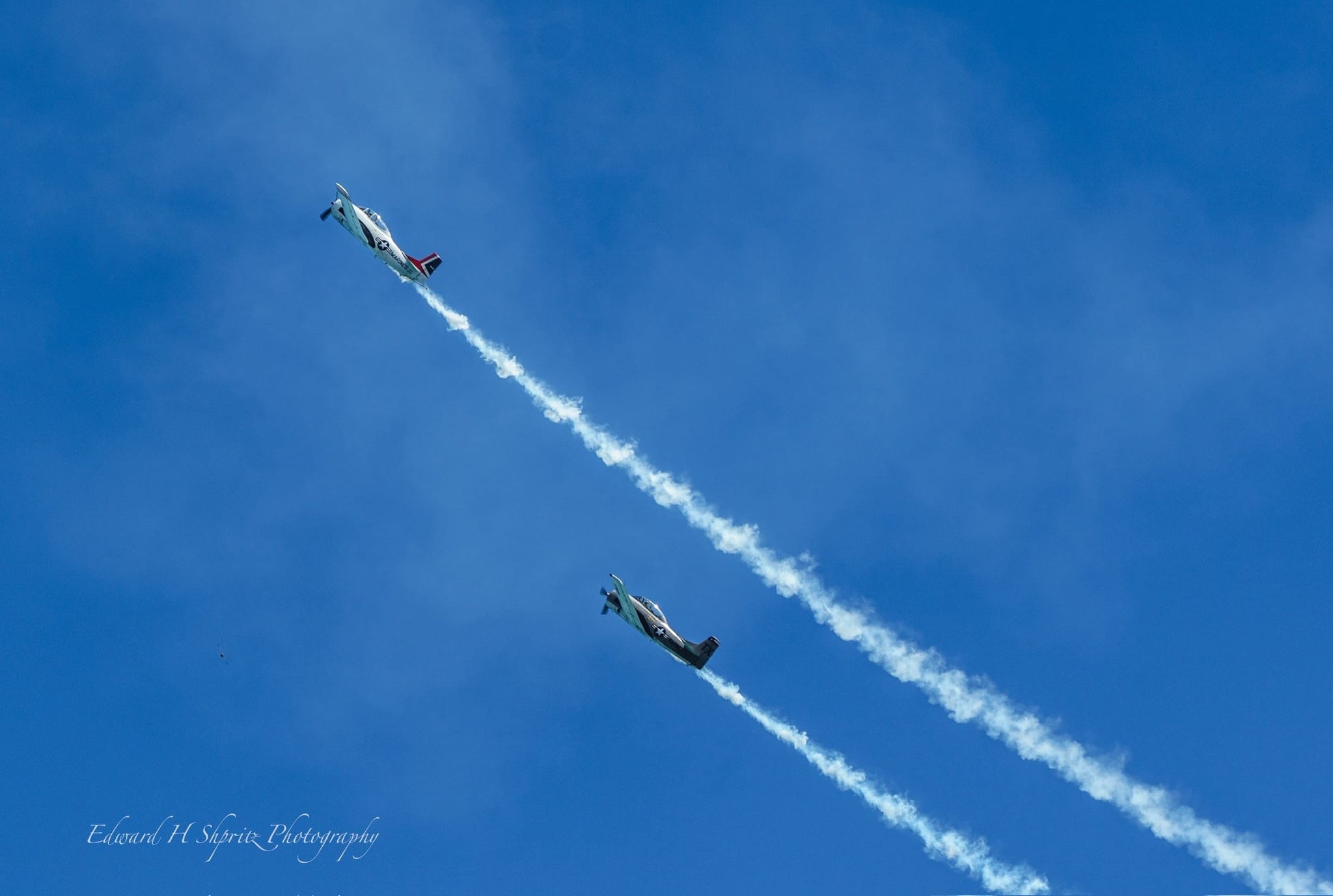 Two aircraft cross the blue sky on separate smoke trails. — by Ed Shpritz