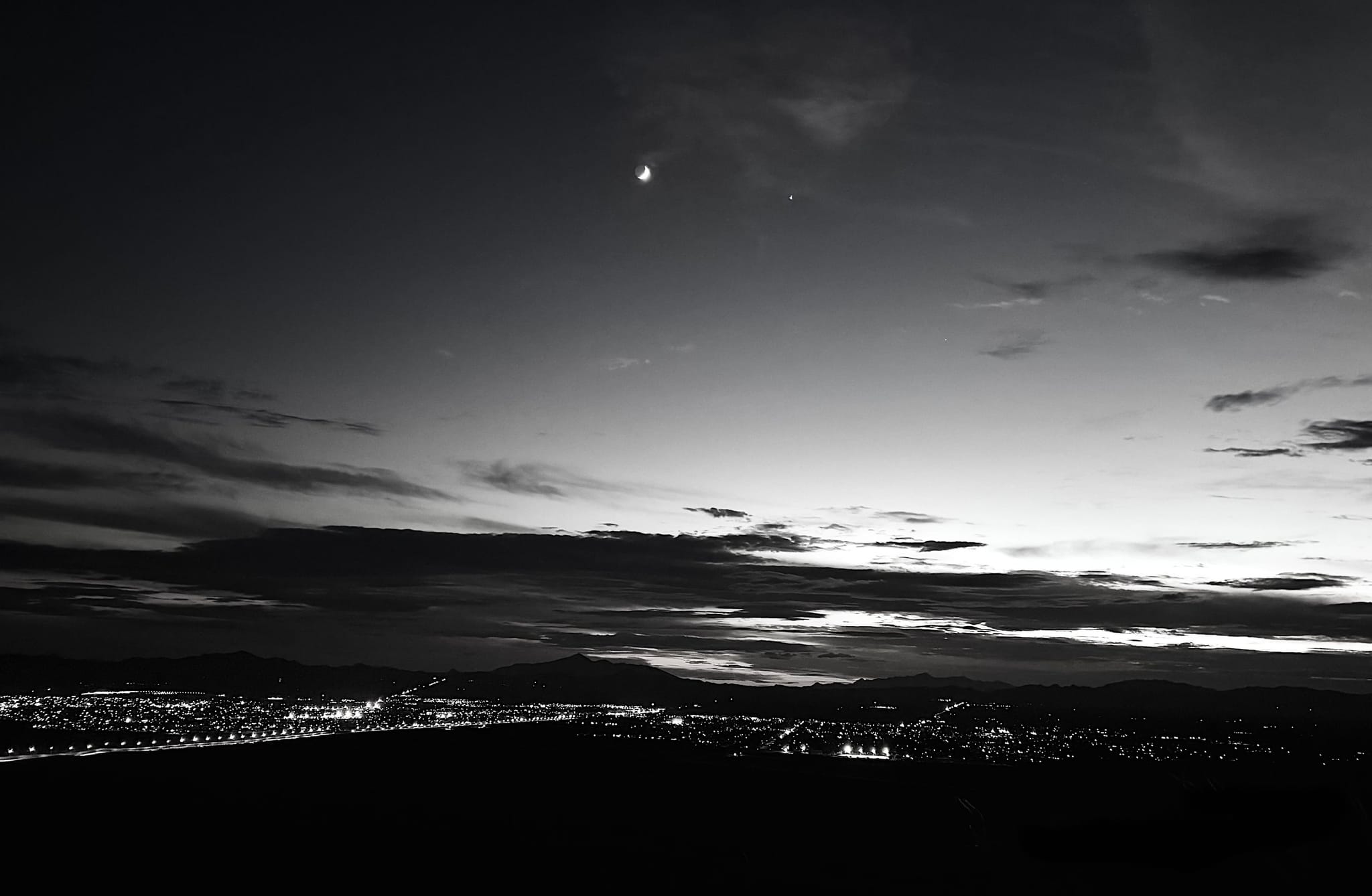 A crescent moon above a distant mountain skyline and city lights at dusk — by Ed Shpritz