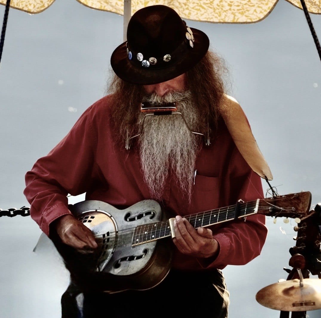 A bearded musician plays a stringed instrument outdoors. — by Ed Shpritz