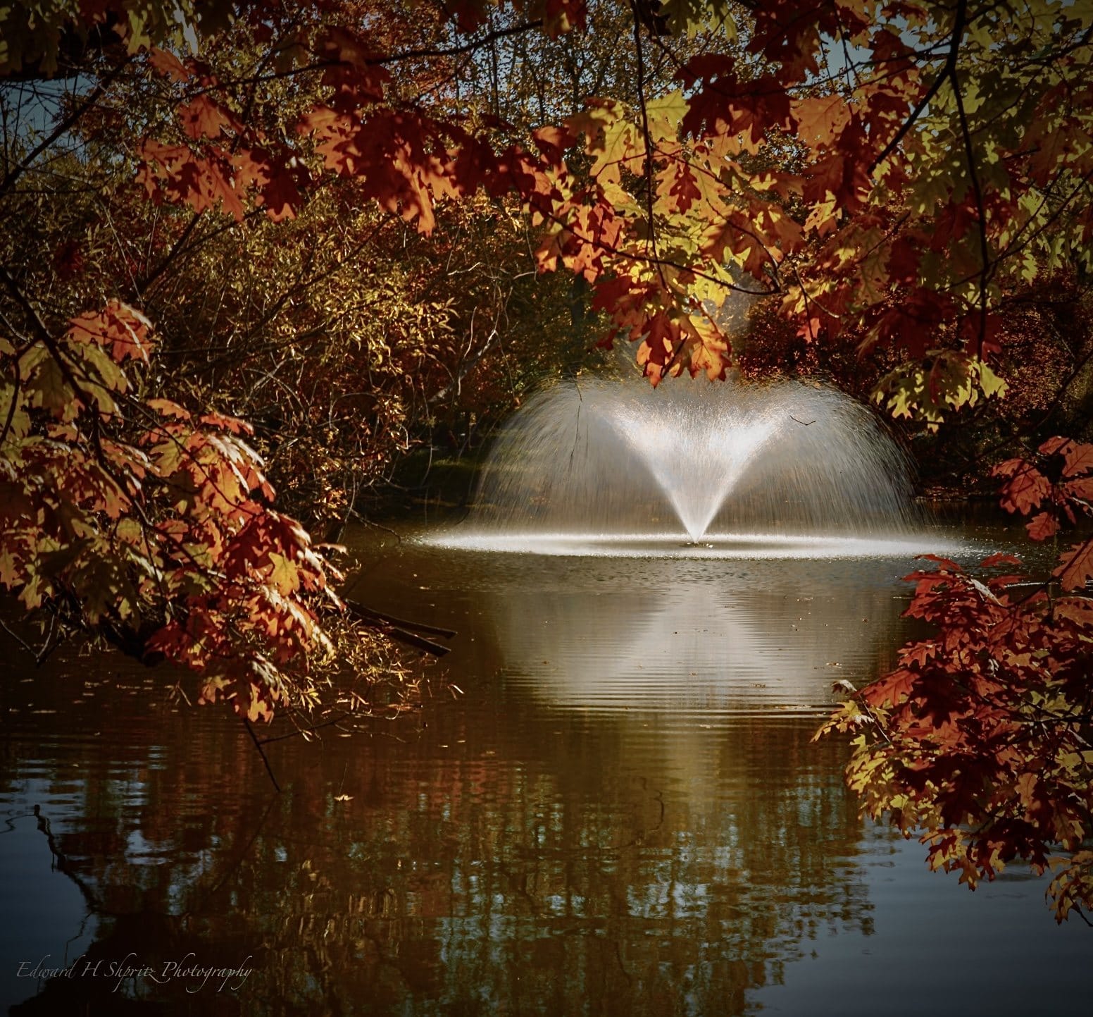 A fountain and its reflection framed by deep red autumn leaves