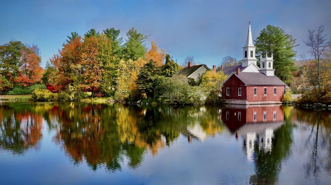 A white New England church surrounded by autumn color and reflected in a still pond