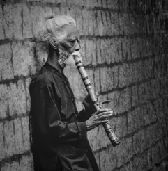 An older man playing a flute against a stone wall, black and white
