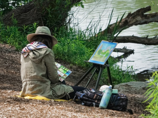 A woman painting at an easel outdoors beneath a large tree